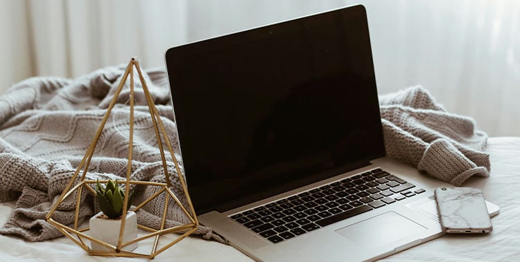 A silver laptop with a black screen sits on a bed covered with a textured gray blanket. To the left of the laptop is a geometric gold wire decor piece containing a small potted succulent plant. To the right of the laptop is a smartphone with a light-colored case. Soft natural light filters through sheer curtains in the background.