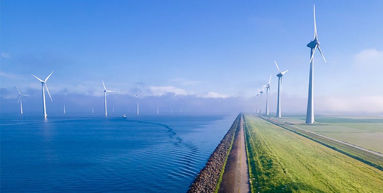 A row of large wind turbines situated along a narrow strip of land that separates a body of water from grassy fields.